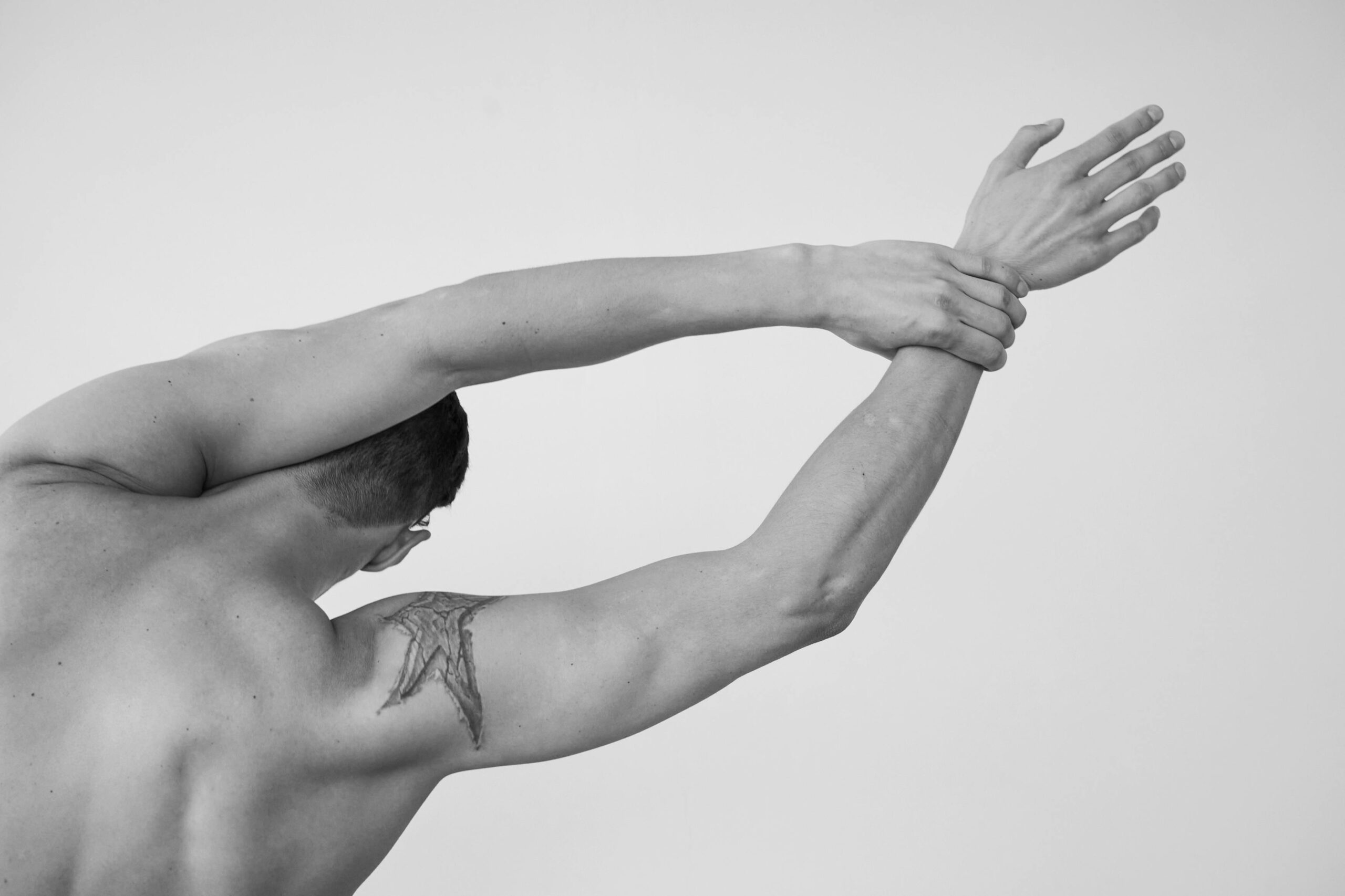 A tattooed man with short hair stretches his arms in a grayscale studio shoot.