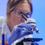 A female scientist wearing goggles and gloves examines a microscope slide in a laboratory.