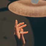 A hand reaches towards a glowing lamp in a warm, dimly-lit indoor setting.