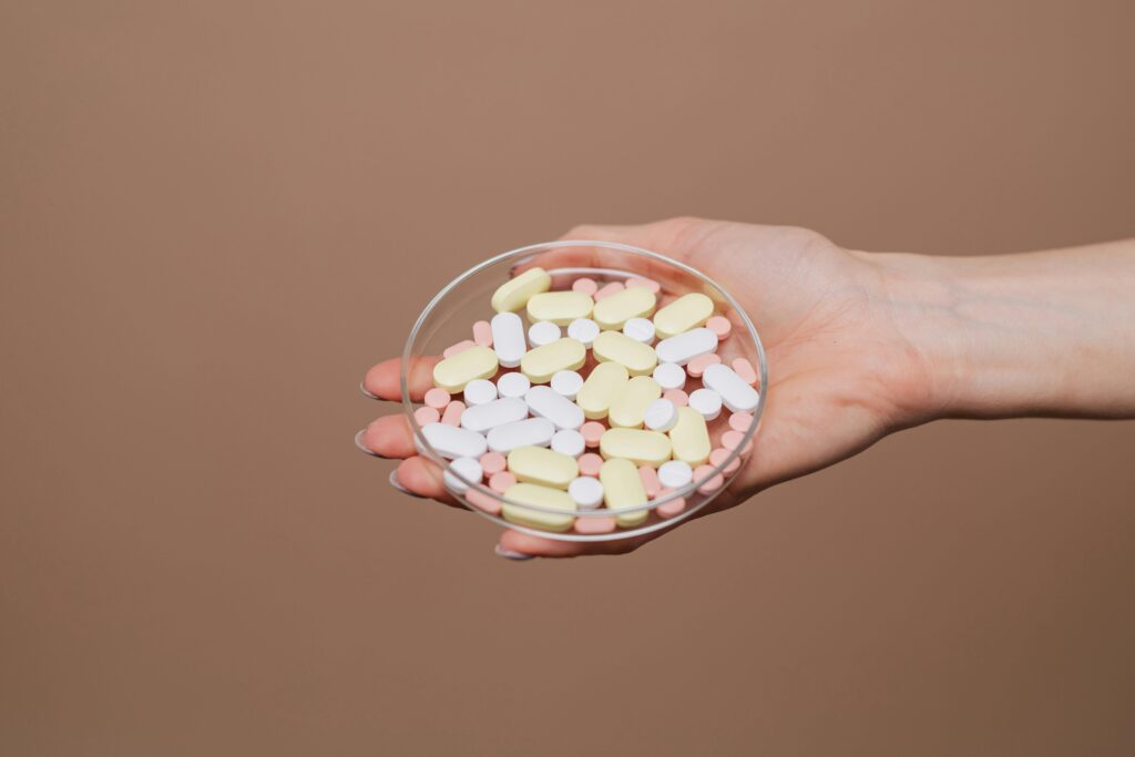 Close-up of a hand holding a petri dish filled with various tablets and capsules on a neutral background.