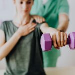 Crop unrecognizable orthopedist in uniform helping slender female in casual wear reaching arm with dumbbell in doctor office in hospital